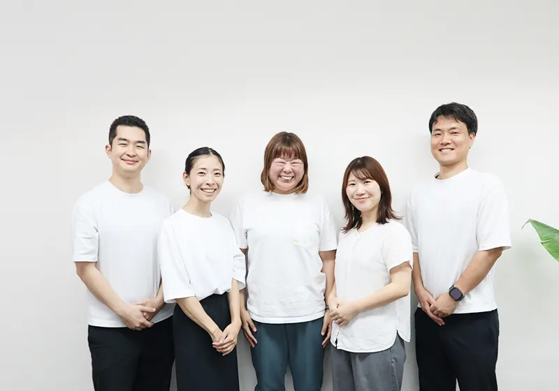白いTシャツを着た5人のスタッフが並んで笑顔で立っている事業所の写真